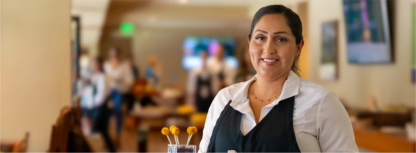 A smiling female Sodexo Live! server holding a tray with desserts and lollipops in a catering or event setting