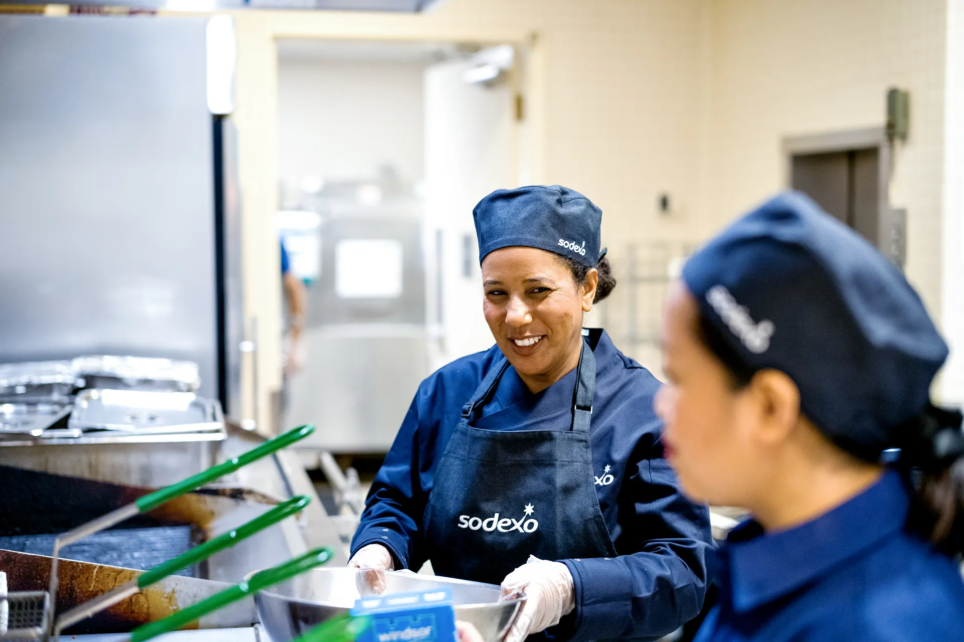 A smiling female Sodexo chef wearing an apron and hat, preparing food in a commercial kitchen