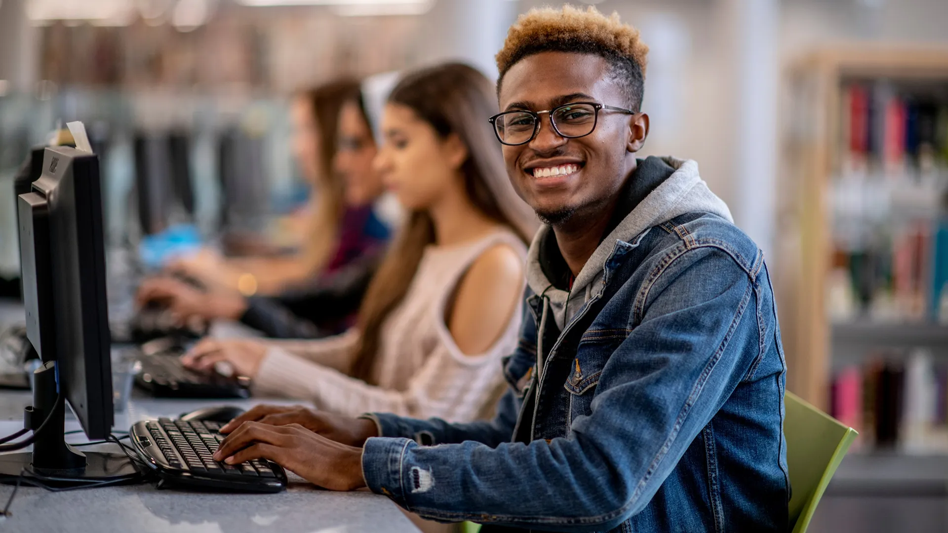 A smiling male student with glasses working on a computer in a library or computer lab, with other students in the background