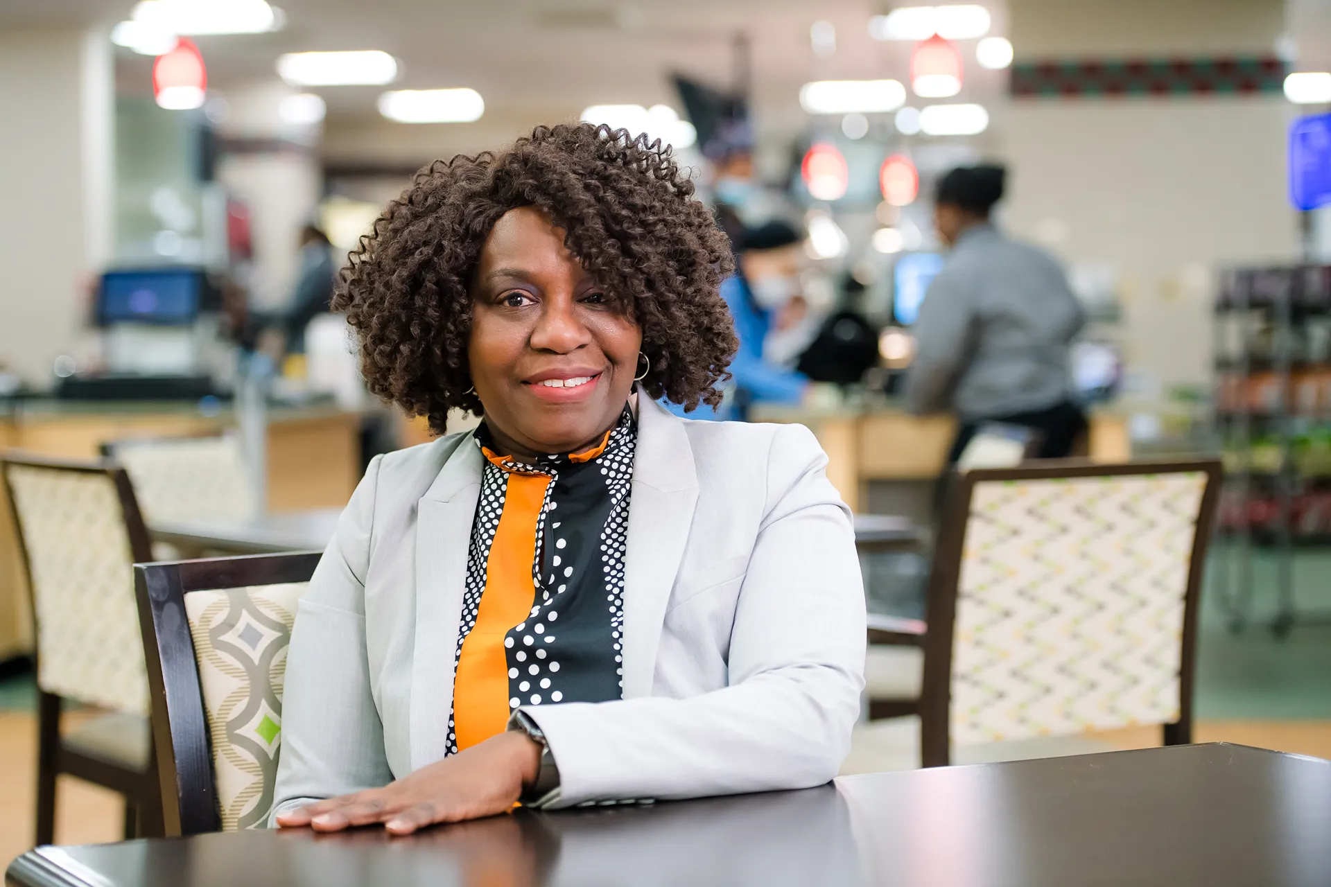A smiling woman with curly hair wearing a grey blazer and patterned blouse, seated at a table in a bright cafeteria or food service area.