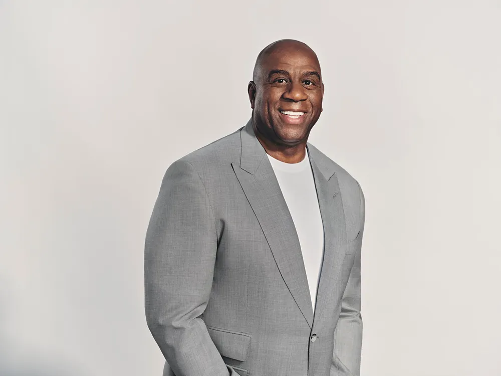Magic Johnson smiling and wearing a light grey suit jacket and white t-shirt against a plain background
