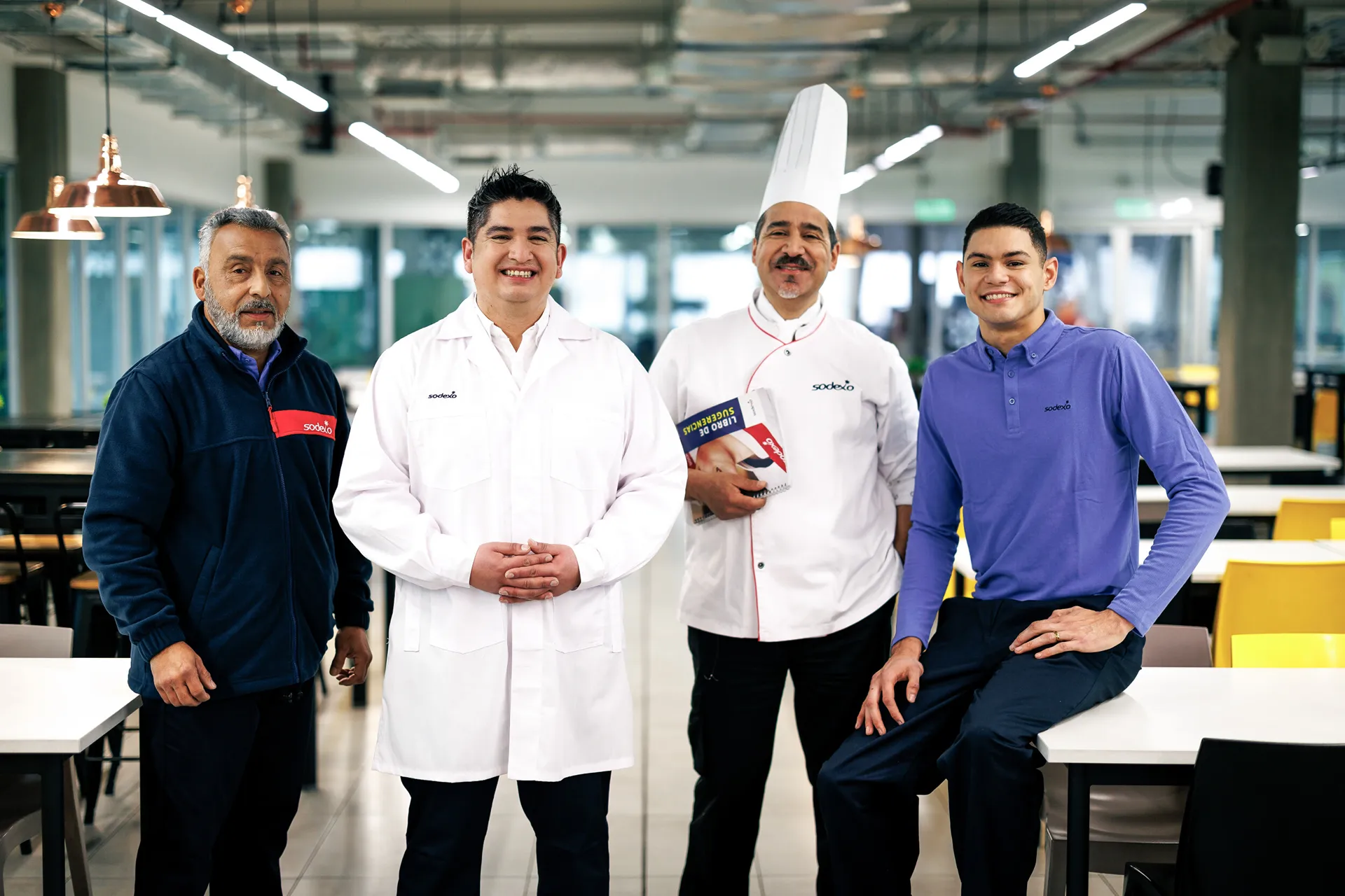 Four smiling male Sodexo employees in various roles, including a chef and a manager, standing in a cafeteria